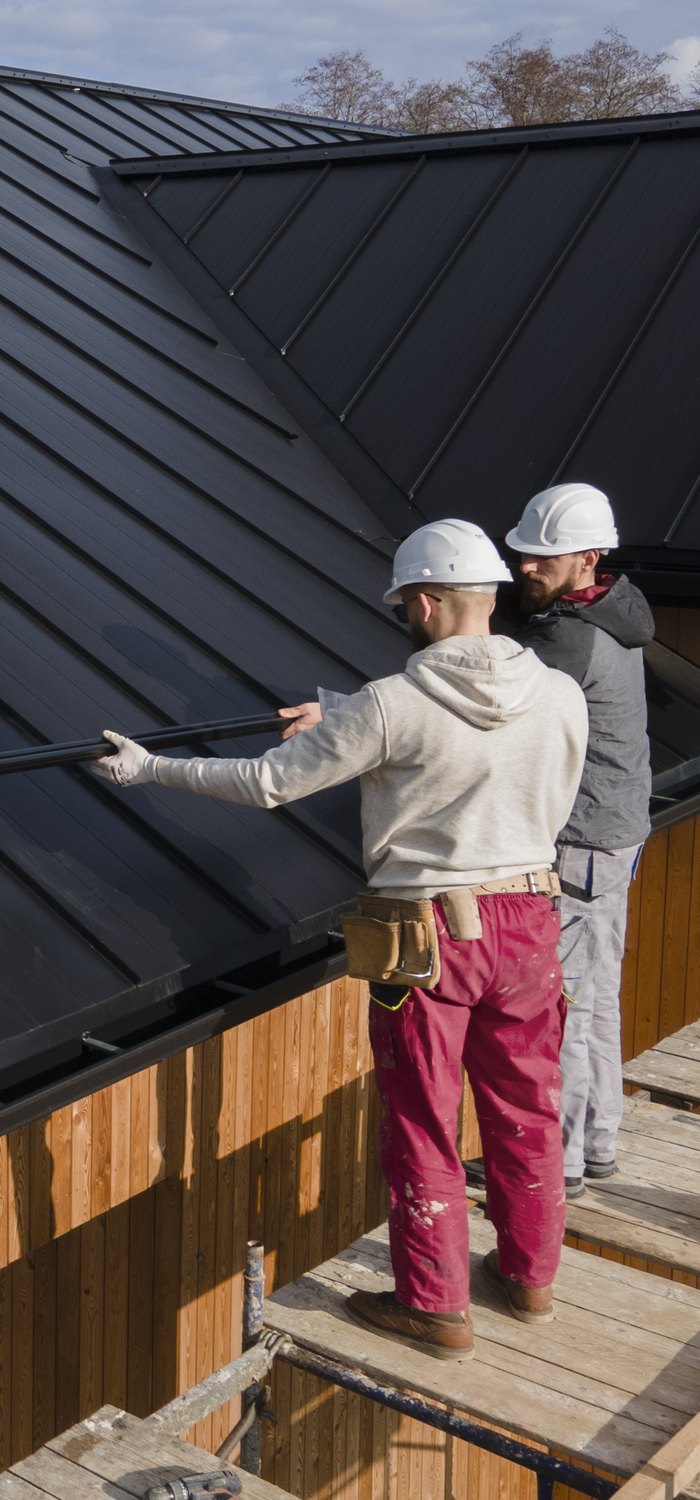 Two roofers fitting a standing-seam panel on a timber-framed house
